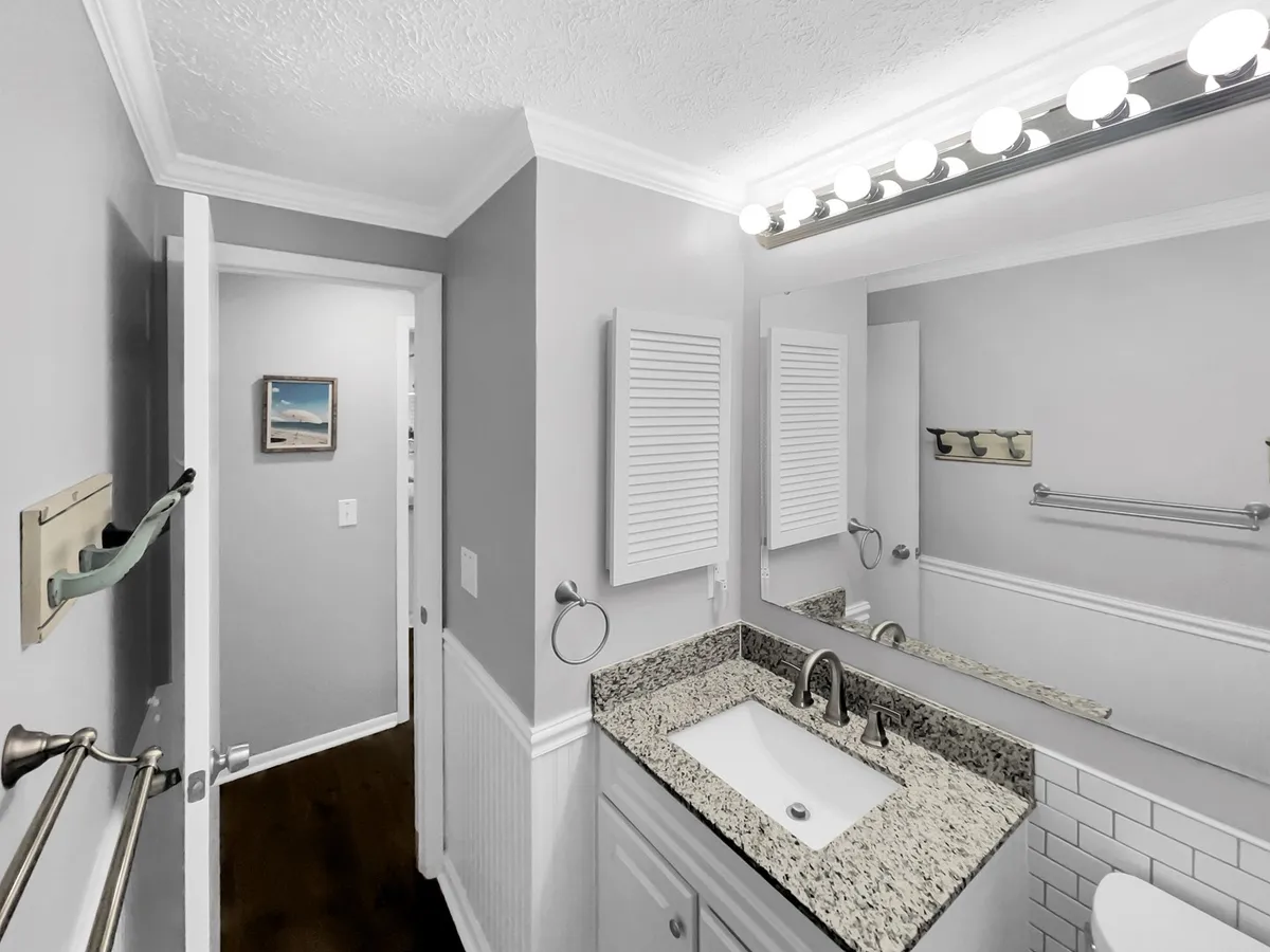 A modern, bright bathroom with granite countertops, a large mirror, contemporary light fixtures, and a glimpse of a glass shower area, reflecting ample space and upgraded finishes.