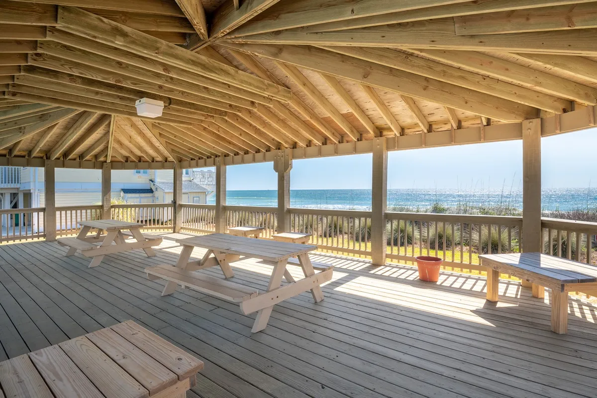 This exterior scene captures wooden picnic tables overlooking the ocean, with an open ceiling design that maximizes the connection to nature. The setup encourages al fresco gatherings in the fresh coastal air, with the sparkling water just beyond.