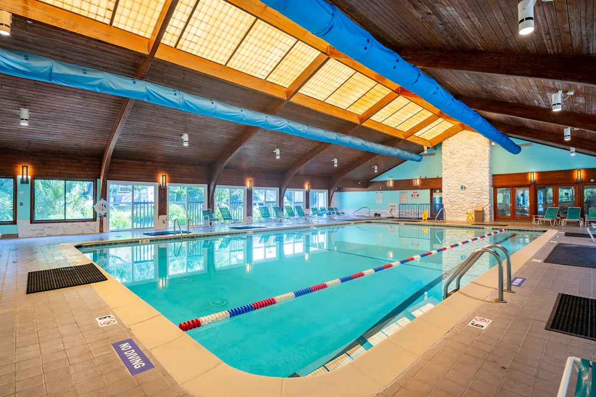 A bright indoor recreation center with a sparkling swimming pool, wooden beams overhead, large windows allowing plenty of daylight, and poolside lounge chairs arranged for relaxation. The atmosphere is cheerful and inviting, suggesting opportunities for swimming, relaxing, and socializing regardless of the weather.