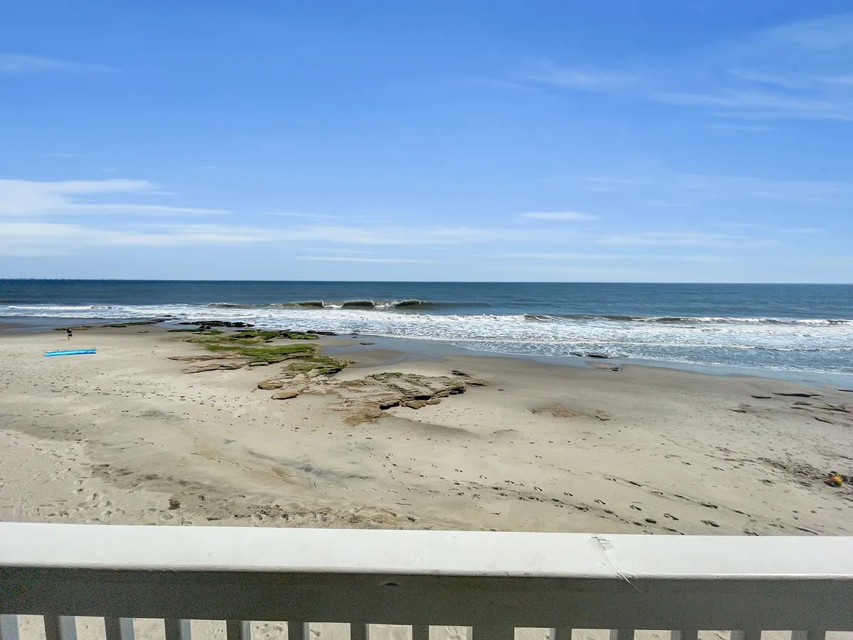 A scenic beach view with rolling ocean waves, rock formations, a surfboard, and a person enjoying the shoreline. The wide expanse of sand and the tranquil seascape invite exploration and adventure.