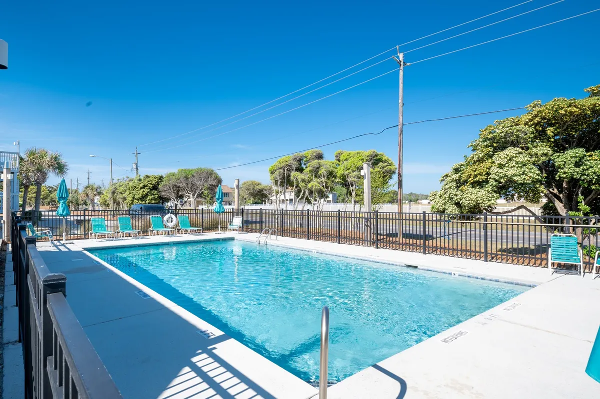 A sparkling outdoor swimming pool surrounded by lounge chairs and lush green trees under a clear blue sky. The pool area exudes a relaxed, vacation-like atmosphere, inviting guests to swim, sunbathe, or socialize in the fresh air.
