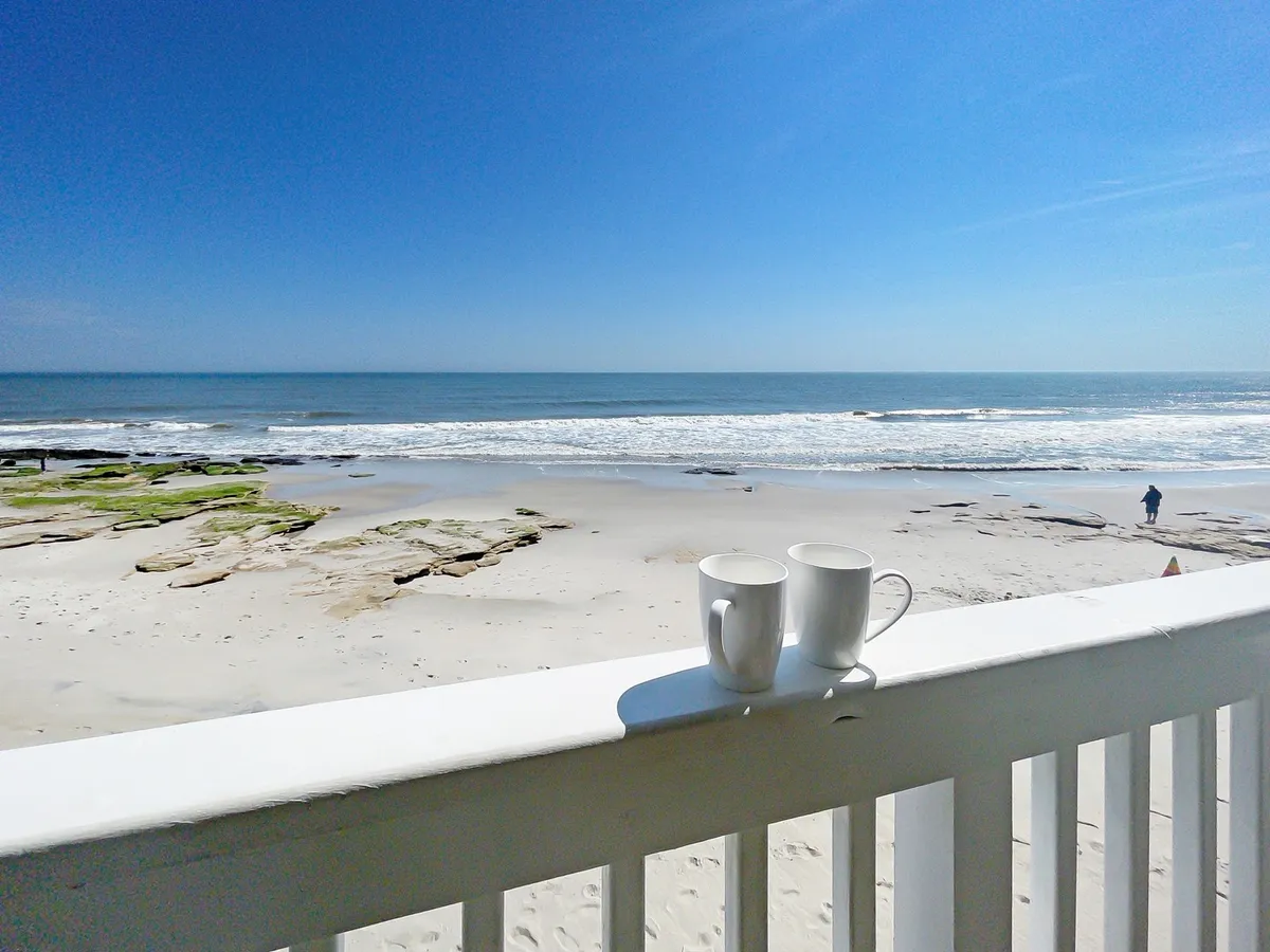 This image shows two white mugs resting on a balcony railing, with a beautiful view of the beach, ocean waves, and natural rock formations in the background. The scene evokes a sense of relaxation and connection to the coastal landscape.