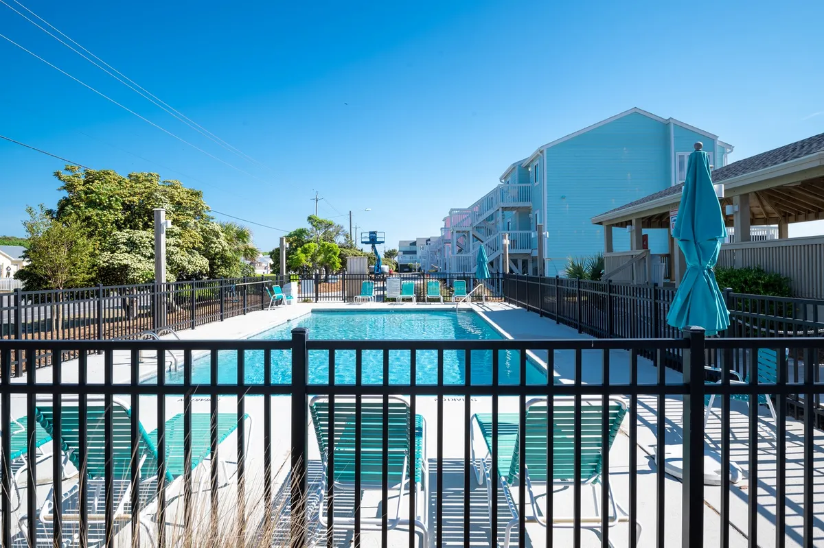 A vibrant outdoor swimming pool scene with lounge chairs and umbrellas arranged by the poolside. The setting is lively yet relaxed, perfect for sunbathing, swimming, or gathering with friends and family. Blue skies and greenery complete the cheerful atmosphere.