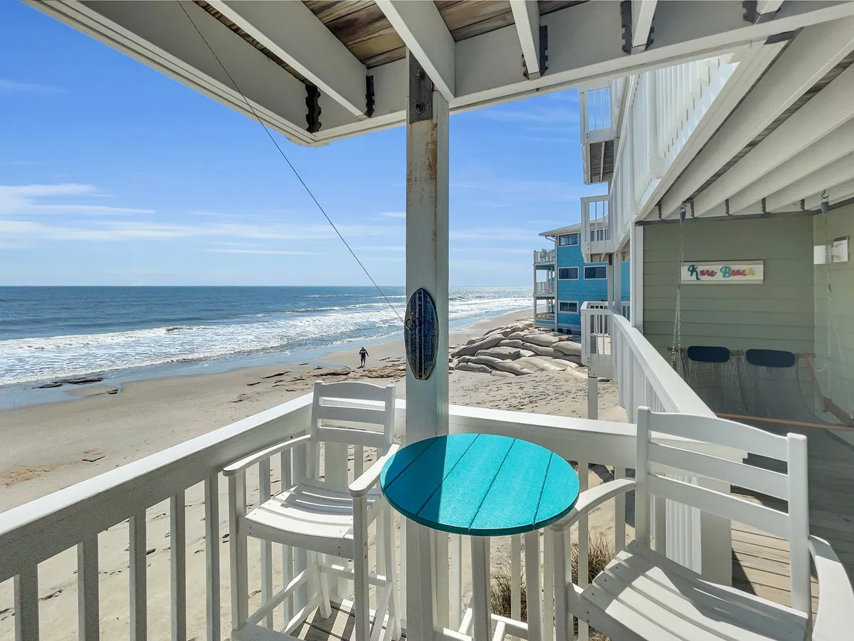 This image captures a tranquil beach scene with waves, blue sky, and inviting outdoor seating including a colorful table. The setting suggests relaxation, direct beach access, and the potential for various beach activities like sunbathing, walking, or reading with an ocean view.