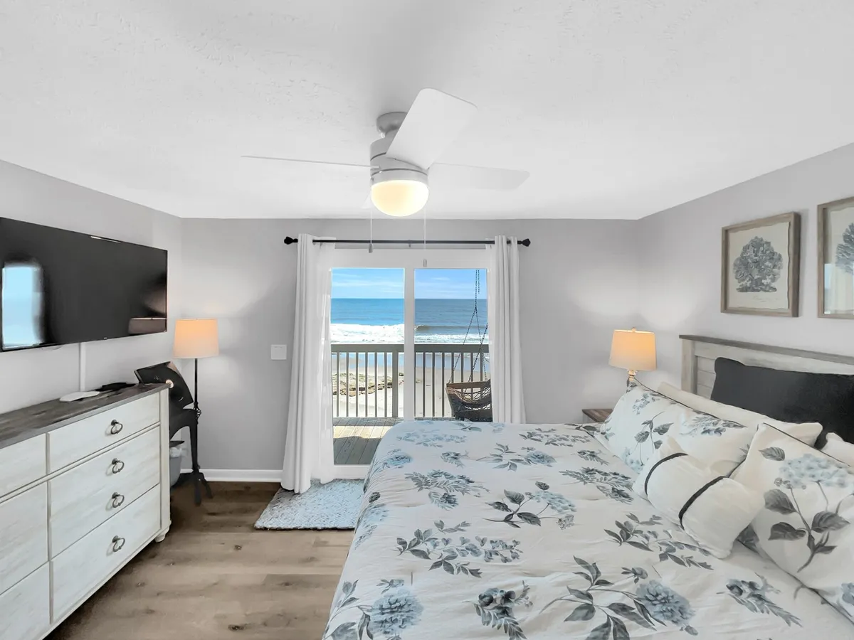 A tranquil bedroom featuring a plush bed with floral bedding, a large window framing sweeping ocean views, a smart TV mounted on the wall, and a decorative lamp on the nightstand. Natural light fills the space, emphasizing comfort and serenity.