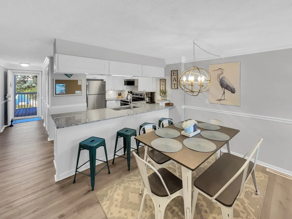 This image showcases a spacious dining area with an elegant table set beneath a sparkling chandelier. The kitchen island is visible in the background, as is an open door leading to a balcony. Decorative art pieces complete the warm and inviting scene, highlighting both communal and culinary spaces.