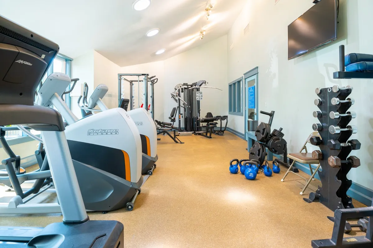 A well-equipped fitness center featuring treadmills, stationary bikes, weight machines, free weights, and exercise mats. The space is bright and organized, with ample room for individual or group workouts.